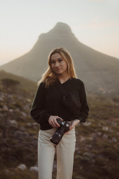 young woman holds a point and shoot camera in her hands with a backdrop of mountains and nature landscape behind her