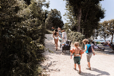 Familie loopt pad op camping omhoog op achtergrond Gardameer voorop vader en zoon daarna moeder en dochter met bolderkar en achteraan twee zoontjes met zwembandjes en zwemvest