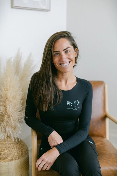 A woman with long brown hair sits on a leather chair, smiling at the camera. She wears a black long-sleeve top and a brown skirt, embodying the confidence and glow often seen at Falmouth Maine Medical Aesthetics, against a neutral background.