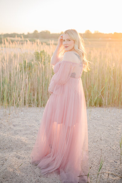 Pregnant mama in a flowing pink gown standing on the beach at sunset, bathed in golden light, capturing a serene and breathtaking moment. Photo by a Connecticut maternity photographer