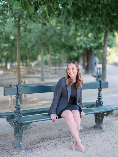 a woman in a dress and blazer sitting on a bench in jardin du luxembourg during the summer