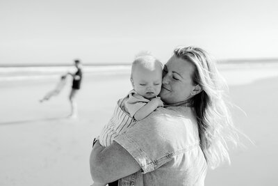 Black and white beach portrait in Geelong showing a mother holding her baby while the father plays with another child in the background by the shoreline.