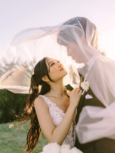 Bride and groom smiling under sunlit veils, holding a bouquet of white roses. 