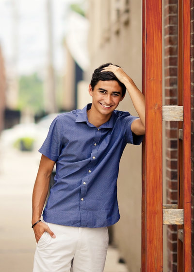 Senior boy leaning against wall with hand in hair, smiling. 