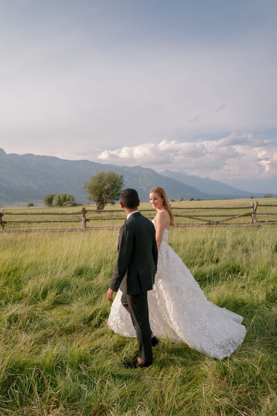 A couple in formal attire walks hand in hand through a grassy field with mountains and a wooden fence in the background.