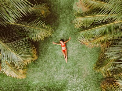 An aerial view of a woman in a vibrant orange bikini lying on lush green grass, surrounded by large, arching palm fronds. This tranquil tropical scene evokes the relaxing moments that vacation fundraising for charities can help create for donors while supporting meaningful causes.