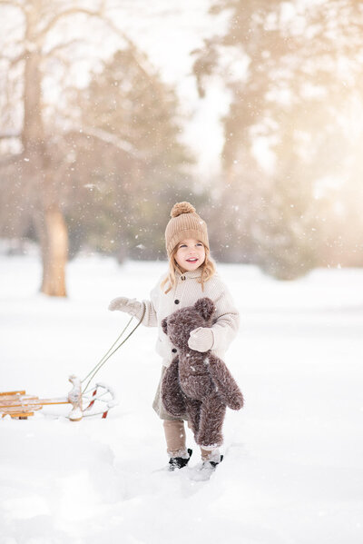 Little girl holding a teddy bear and pulling a wooden sled through the snow in Kamloops. Portrait by Brittany Danielle.