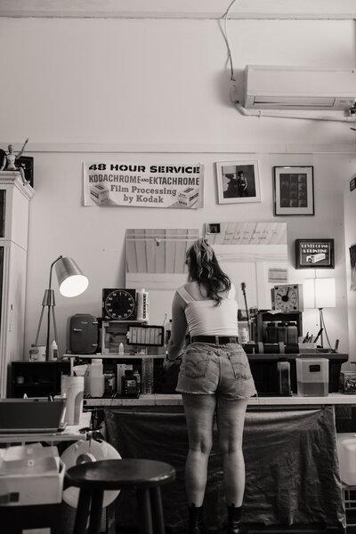 A black-and-white film photograph from a film lab branding session, capturing a lab technician standing at a workstation surrounded by vintage film processing equipment. The nostalgic setting features a retro Kodak sign, framed photographs, and a classic darkroom timer, evoking a timeless appreciation for analog photography. The image highlights the hands-on craftsmanship and authentic charm of the film lab's process.