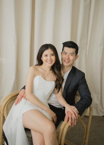 Kohnur Elegant bride in a white strapless dress with soft glam makeup and loose waves sits smiling next to her groom in a modern, light-filled engagement portrait.