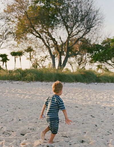 A young boy in a striped outfit walks on a sandy beach toward green grass and trees under a clear sky, evoking a peaceful and playful mood.