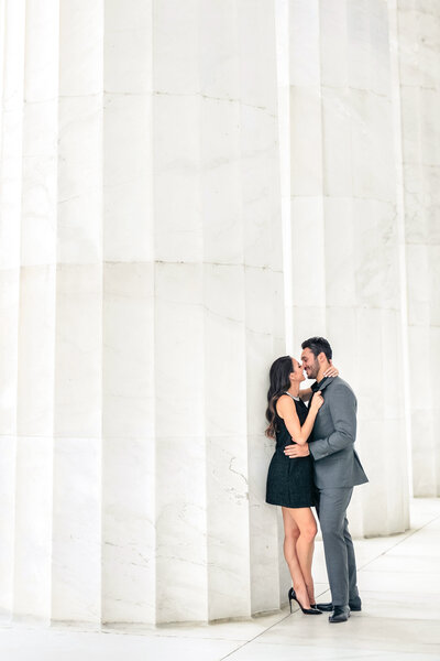 A couple stands closely together, smiling and embracing against a large, white, textured wall. The woman is in a short black dress and heels, while the man wears a gray suit. The setting is bright and airy, with a minimalist architectural background.