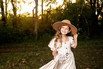 Little girl in a white and tan striped dress wearing an oversized tan hat. She is swaying her dress back and forth in front of the sunlight and greenery.