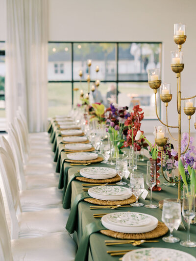 A colorful photograph of a head table at a wedding reception featuring green linens, patterned plates, and eclectic florals.