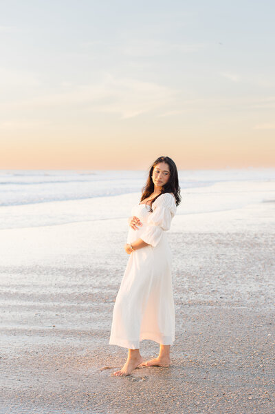 Orlando maternity photographer captures mom standing on the beach at sunrise holding her bump for pregnancy photos