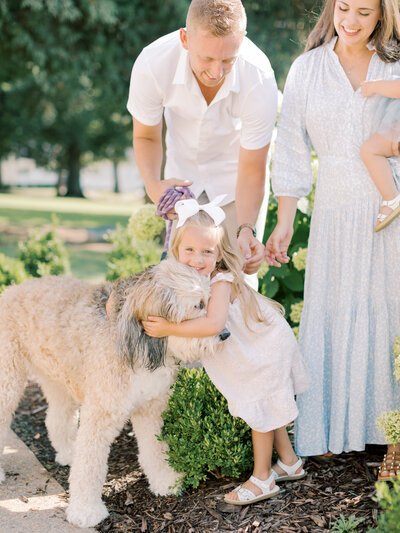 Young, blonde girl in blush pink dress smiles while hugging large dog while standing in front of mother in blue dress, dad in white shirt taken by Little Rock family photographer Bailey Feeler Photography