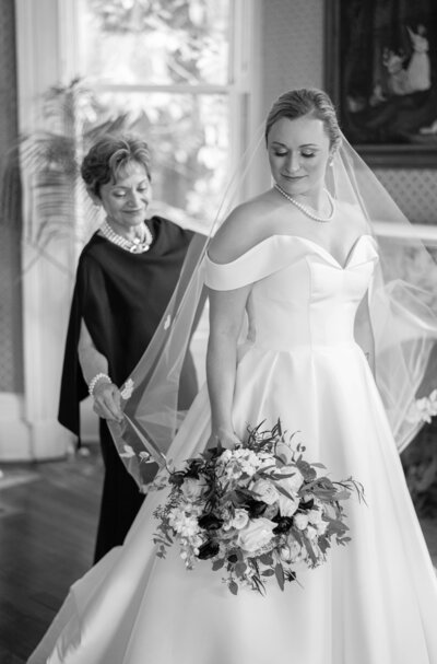 A bride in an off-shoulder gown holds a bouquet, smiling as an older woman adjusts her veil. They stand in a room with elegant decor, and a window with natural light shining through in the background.