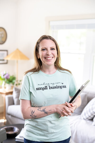 A smiling woman with light brown hair holds a tablet while wearing a mint-green t-shirt that reads "minding my own small business." She stands in a bright, cozy living space with natural light.