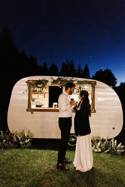 Bride and groom standing in front of vintage mobile bar at nighttime, bride wears grooms jacket at Dallenbach Ranch wedding, Colorado.