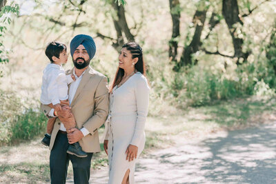 Dad holding son and looking at camera. Mom standing beside dad and looking at son. 