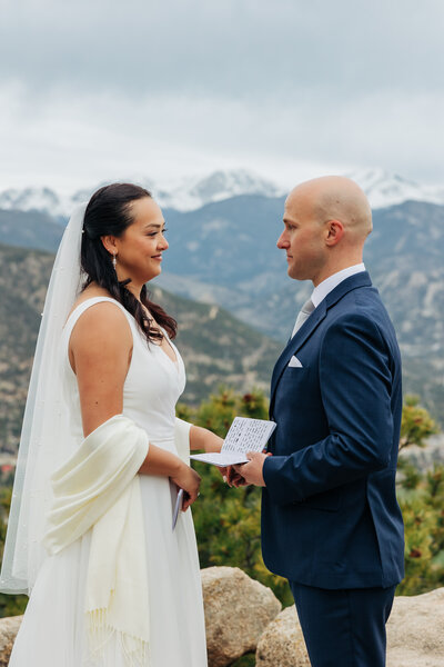the bride and groom are sharing vows together and looking into each other eyes. They're holding hands and the groom has a vow book in his left hand. the bride has a veil and a shawl. There are snowy mountain peaks in the background