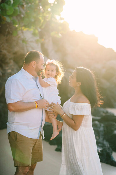 mom and dad holding a toddler on a beach with the sun shining