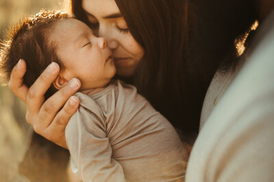 Mother leans in to kiss her newborn baby at sunset.