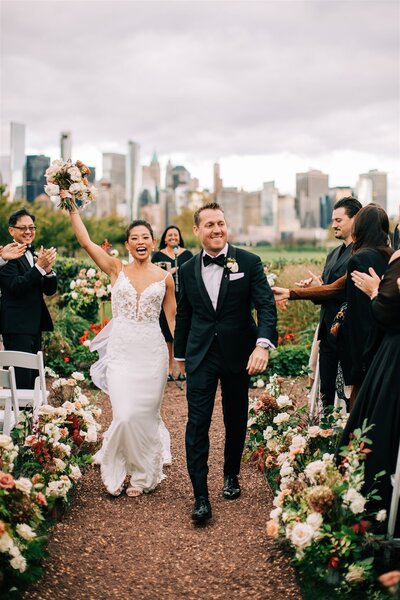 Bride and groom walking down the aisle outdoors with a city skyline in the background, celebrating their wedding with guests cheering.