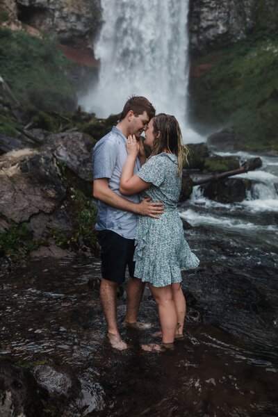 A couple splashes with water stands facing each other with his arms around her waist and her arms on his chin as they face each other with Tumalo Falls flowing behind them for their Oregon adventure engagement and proposal session near Bend. | Erica Swantek Photography