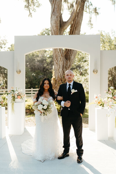 Bride and groom standing in front of the white ceremony arch at Uliana Estate in Lakeland, Florida, surrounded by floral arrangements and oak trees