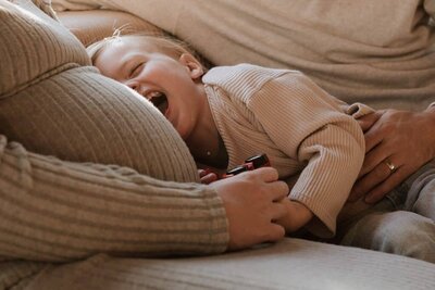 Cozy maternity photo capturing a joyful toddler resting their head on a pregnant belly, smiling with laughter in a warm, intimate family moment.