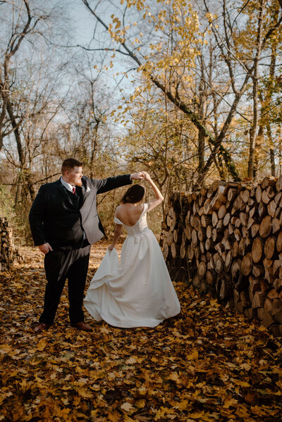 wedding couple twirling with orange and yellow fall leaves on ground next to wall of chopped wood