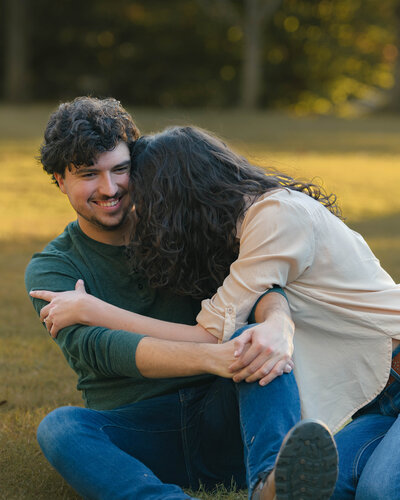 A man and woman sit on some grass at Centennial park in Nashville, TN hugging and laughing.