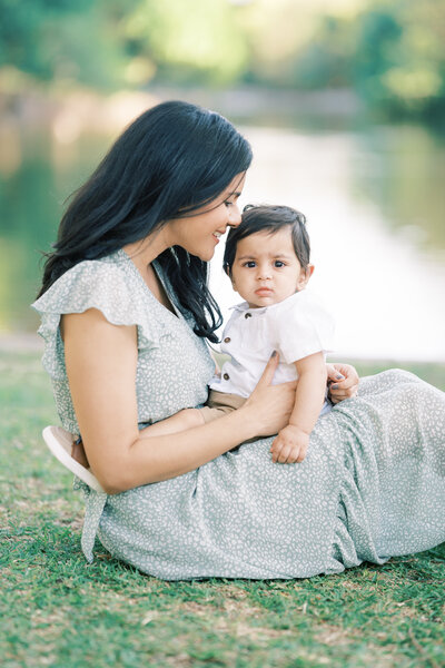 Portrait of a woman wearing a blue sundress while sitting smiling at a baby boy sitting on her lap in front of a lake.