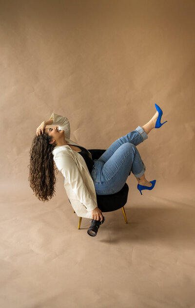 A portrait of Hiba sitting on a small black chair. She has her feet in the air with one hand in her hair and the other holding her camera.