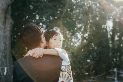 木漏れ日の中で父親に抱きつく男の子の笑顔、家族の絆を感じる七五三の風景
