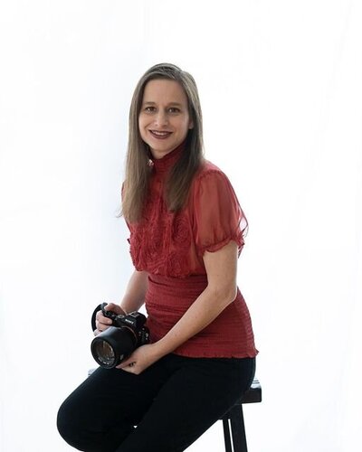 Portrait of a boudoir studio owner in San Fransisco, seated on a stool with a warm smile, holding her camera, dressed in a red shirt.