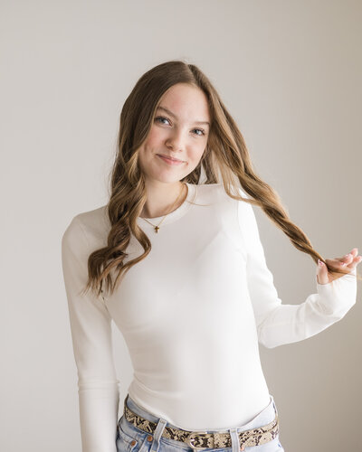 high school senior female, standing in a studio, with long dark hair, playing with some of her hair, facing the camera and smiling, wearing a long sleeve white shirt.