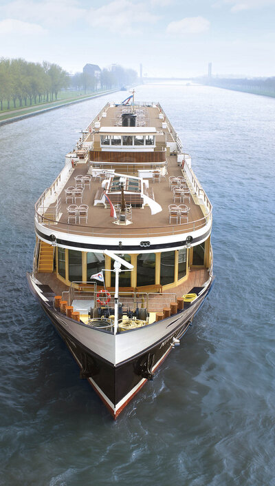 A large river cruise ship sails forward along a wide, calm waterway, with multiple open decks, seating areas, and railings visible; trees line the distant riverbank under a lightly cloudy sky.
