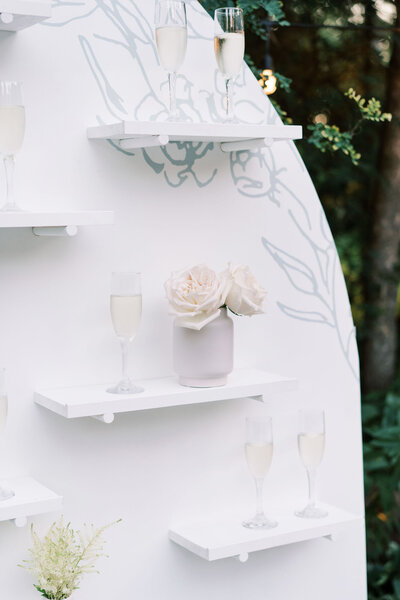 A white circular shelf, designed by a talented wedding planner in Calgary, holds several champagne glasses and a vase with light pink roses, set against a backdrop of greenery.