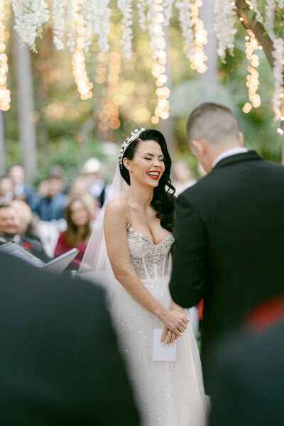 bride crying and smiling as groom reads his vows  standing under dripping flowers and twinkle lights at hartley botanica in somis captured by magnolia west photography los angeles wedding photographers