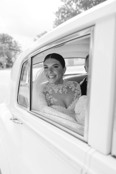 Bride smiling, captured through the window of a classic white wedding car.