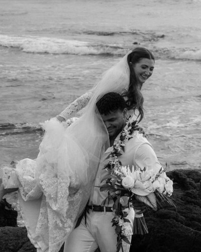 Black and white image of a groom carrying his bride, capturing a timeless and romantic moment from their luxury destination wedding