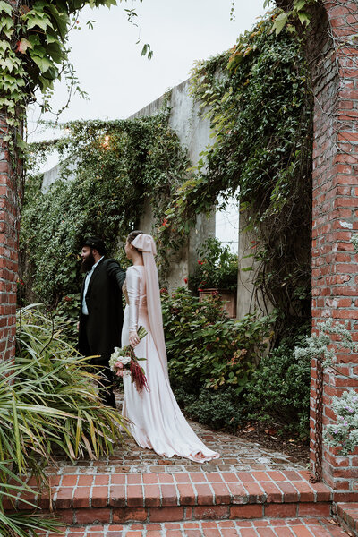 Editorial-style hasidic wedding portrait of a bride and groom walking through a lush, overgrown garden ruin. The bride wears a long-sleeved gown with a veil and holds a bouquet, creating a timeless, romantic atmosphere.