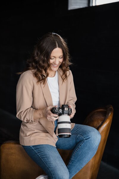 A person with long hair, wearing a beige blazer and jeans, sits on a brown chair and looks at a camera they're holding—perhaps capturing inspiration for their photography website design.