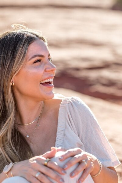 Natalie Bernacchi laughing in the sunlight, holding a cup, with red rock landscape in the background—radiating joy and ease
