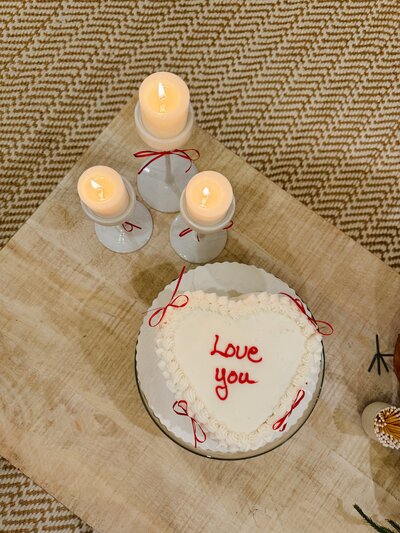 Heart-shaped cake with "Love you" in red icing on a wooden table, surrounded by white candles with red ribbons, creating a cozy, romantic ambiance.