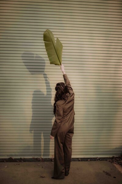 A woman in a brown suit stands with her back to the camera, holding a large green leaf overhead as her shadow casts dramatically on a pale green corrugated wall in warm afternoon light.