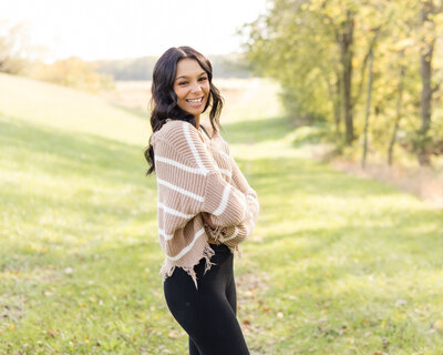 High school senior female with dark brown hair and eyes, wearing a light tan and white stripped shirt with black leggings, smiling at the camera