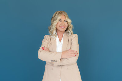 Divorce coach standing confidently with crossed arms in front of a blue background, dressed in a beautiful outfit. This image symbolizes strength, resilience, and the beginning of a new chapter after divorce.