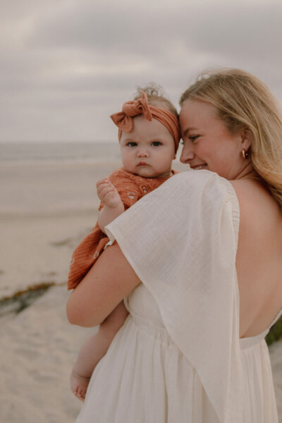 Mother holding her baby wrapped in a rust-colored outfit by the beach